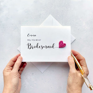Personalised bridal party proposal cards. White card with black cursive script text. The card at the front reads 'Emma, will you be my Bridesmaid' there is a bright pink crochet heart at the end of the 'd' - adding a pop of colour. 