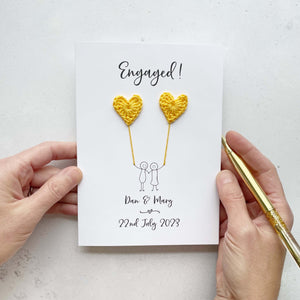 A white card featuring a line drawn couple each holding a yellow crochet heart balloon. Engaged! is printed at the top in black cursive text. The couples names and date of engagement is printed at the bottom.