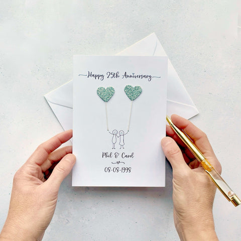 A white card featuring a line drawn couple holding hands and each holding a heart shaped balloon which is made from sparkly silver coloured card. Happy 25th Anniversary is printed at the top of the card and the couples names and wedding date are printed at the bottom. 