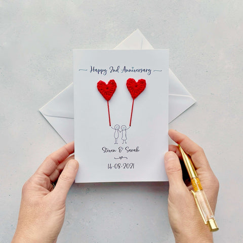 A white card featuring a line drawn couple each holding a red crochet heart made from cotton yarn. Happy 2nd Anniversary is printed at the top of the card and the couples names and wedding date are printed at the bottom. 