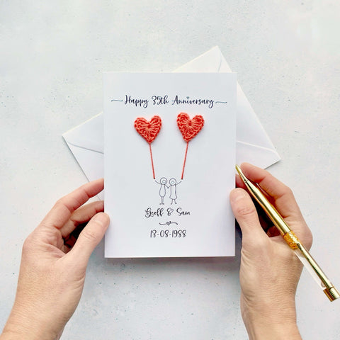 A white card featuring a line drawn couple holding hands and each holding a crochet heart shaped balloon which is made using coral coloured cotton yarn. Happy 35th Anniversary is printed at the top of the card and the couples names and wedding date are printed at the bottom. 