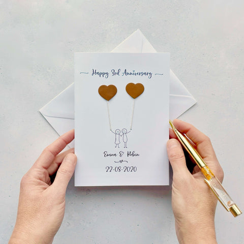 A white card featuring a line drawn couple each holding a leather heart shaped balloon. Happy 3rd Anniversary is printed at the top of the card and the couples names and wedding date are printed at the bottom. 