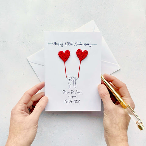 A white card featuring a line drawn couple holding hands and each holding a crochet heart shaped balloon which is made using red coloured cotton yarn. Happy 40th Anniversary is printed at the top of the card and the couples names and wedding date are printed at the bottom. 