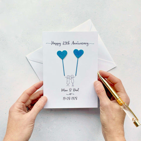 A white card featuring a line drawn couple holding hands and each holding a heart shaped balloon which is made using sparkly blue card. Happy 45th Anniversary is printed at the top of the card and the couples names and wedding date are printed at the bottom. 