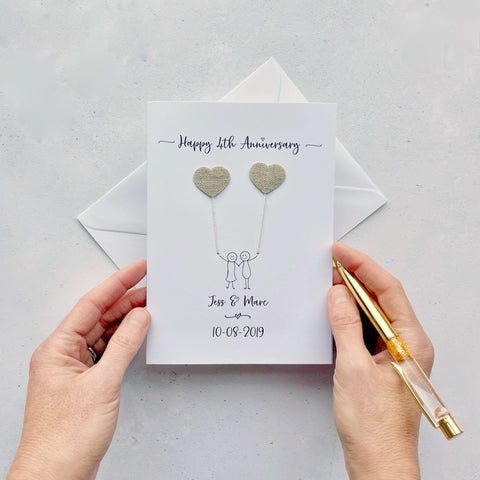 A white card featuring a line drawn couple each holding a heart shaped balloon which is made from linen. Happy 4th Anniversary is printed at the top of the card and the couples names and wedding date are printed at the bottom. 