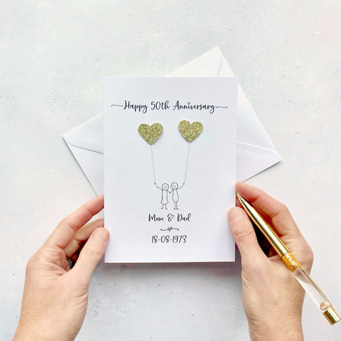 A white card featuring a line drawn couple holding hands and each holding a heart shaped balloon which is made using sparkly gold card. Happy 50th Anniversary is printed at the top of the card and the couples names and wedding date are printed at the bottom. 