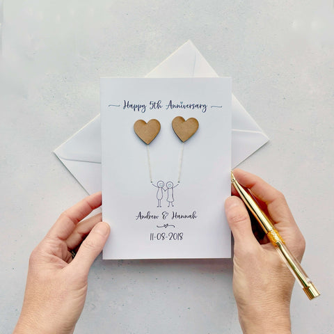 A white card featuring a line drawn couple holding hands and each holding a heart shaped balloon which is made from MDF. Happy 5th Anniversary is printed at the top of the card and the couples names and wedding date are printed at the bottom.