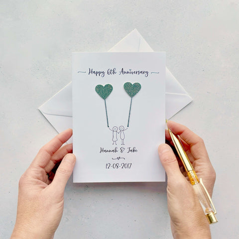 A white card featuring a line drawn couple each holding a iron coloured sparkly heart made from cardboard. Happy 6th Anniversary is printed at the top of the card and the couples names and wedding date are printed at the bottom. 