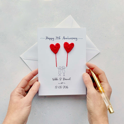 A white card featuring a line drawn couple holding hands and each holding a heart shaped balloon which is crocheted using wool yarn. Happy 7th Anniversary is printed at the top of the card and the couples names and wedding date are printed at the bottom. 