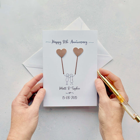 A white card featuring a line drawn couple holding hands and each holding a heart shaped balloon which is made from bronze coloured card. Happy 8th Anniversary is printed at the top of the card and the couples names and wedding date are printed at the bottom. 
