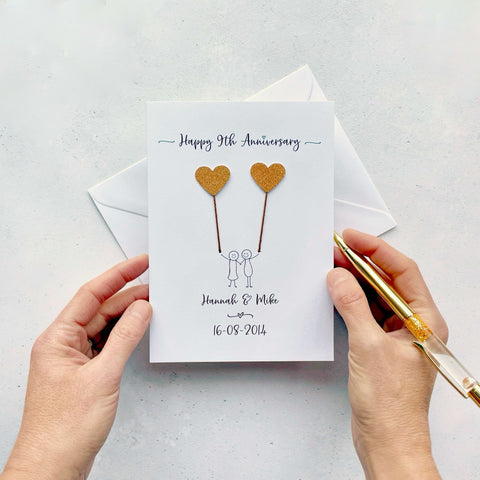 A white card featuring a line drawn couple holding hands and each holding a heart shaped balloon which is made from sparkly copper coloured card. Happy 9th Anniversary is printed at the top of the card and the couples names and wedding date are printed at the bottom. 