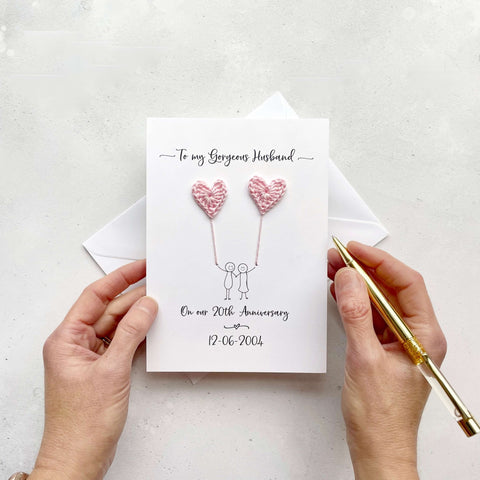 A white card featuring a line drawn couple holding hands and each holding a pale pink crochet heart balloon. ‘To my Gorgeous Husband’ is printed at the top of the card and at the bottom of the card we have printed: ‘On our 20th anniversary’ and their wedding date. 