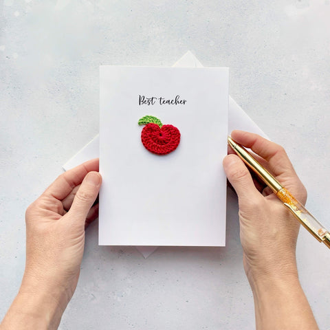  A white card with a red crochet apple attached to the card. Above the apple it reads ‘Best Teacher’ in black cursive text. If you choose to personalsie this design, the teachers name will be printed underneath the apple. 