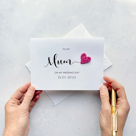 A white card with 'to my Mum on my wedding day' printed in black text. The wedding date is also on there too. The word 'Mum' is much larger than the rest of the text. There is a bright pink crochet heart coming off the 'm' in 'Mum'.