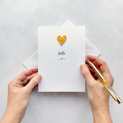 A white card with a cute little yellow handmade crochet heart stuck in the centre, the word ‘Hello‘ has been printed below the heart in black cursive text. 