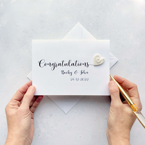 A white card with 'Congratulations' printed across the card in large black script text. There is a white crochet heart at the end of that word and underneath 'Congratulations', you've got the two names and their wedding date. 