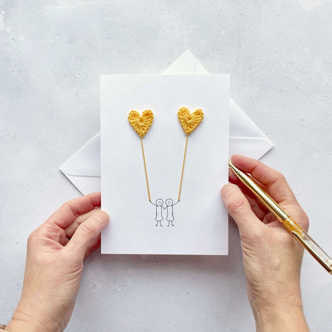 A white card featuring a line drawn female couple holding hands and each holding a yellow crochet heart balloon. 