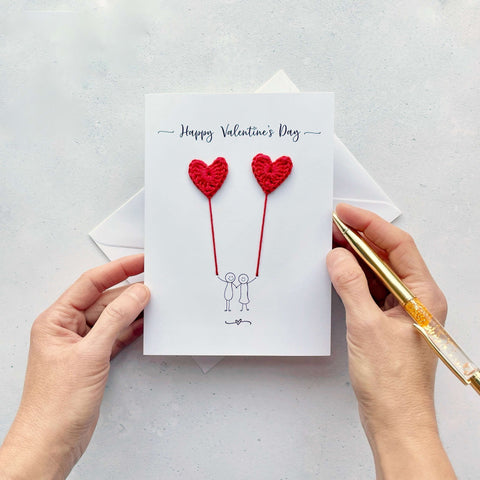 A white card featuring a line drawn couple holding hands and each holding a red crochet heart balloon.  ‘Happy Valentine’s Day’ is printed at the top of the card in black cursive text.