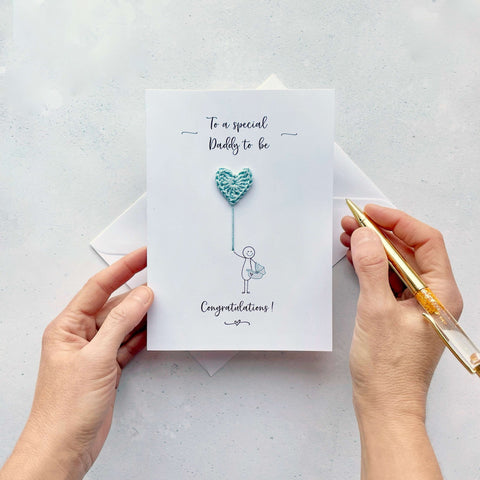A card featuring a line drawn male, holding a pale blue crochet heart balloon. ‘To a special Daddy to be’ is printed at the top of the card in black cursive text .The character is drawn holding a baby car carrier and the word ‘Congratulations!’ is printed below him.