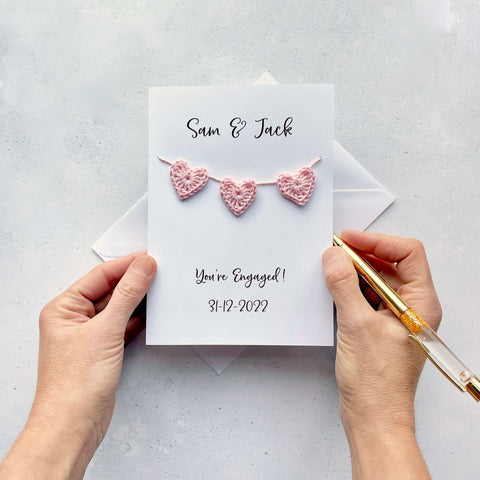 A white greetings card with two names printed at the top of the card in black cursive text. Just under the names there are 3 mini crochet hearts strung across the card. The words 'You're Engaged!' and the date 31-12-2022 are printed at the bottom of the card. 