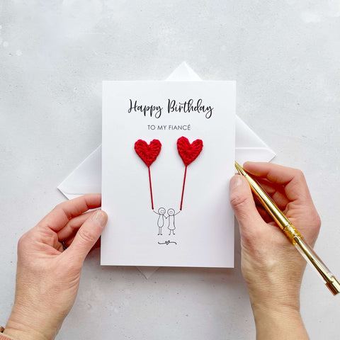 A white birthday card featuring a line drawn couple holding hands and each holding a red crochet heart balloon.  ‘Happy Birthday to my Fiancé’’ is printed at the top of the card in black cursive text.