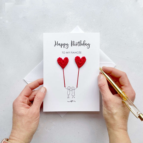 A white birthday card featuring a line drawn couple holding hands and each holding a red crochet heart balloon.  ‘Happy Birthday to my Fiancée’’ is printed at the top of the card in black cursive text.
