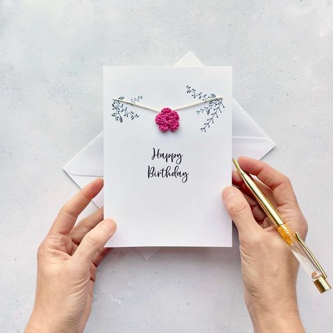 A white card with 'Happy Birthday' printed in the centre of the card in black cursive text. There is some line drawn foliage avove this wording and a bright pink crochet flower has been strung across the card on white yarn.