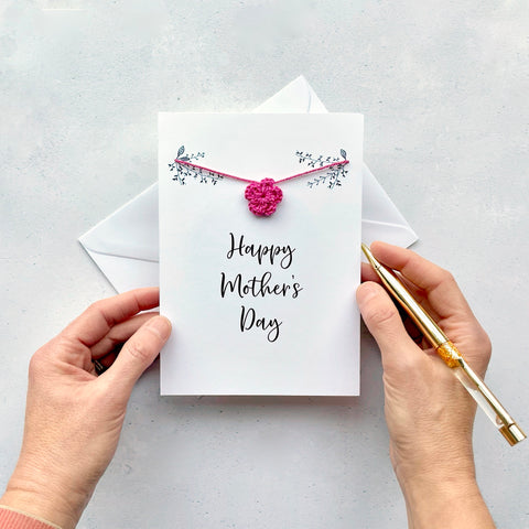 A white Mother's Day card with a bright piink crochet flower strung across the front.  Line drawn foliage has been printed on eachh side of the card in black and the words 'Happy Mother's Day has also been printed in black (Cursive text) 