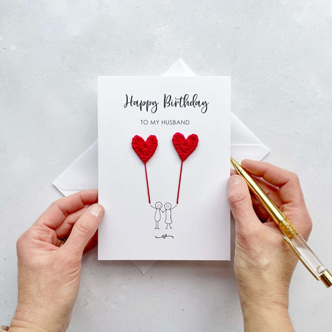 A white birthday card featuring a line drawn couple holding hands and each holding a red crochet heart balloon.  ‘Happy Birthday to my Husband’ is printed at the top of the card in black cursive text.
