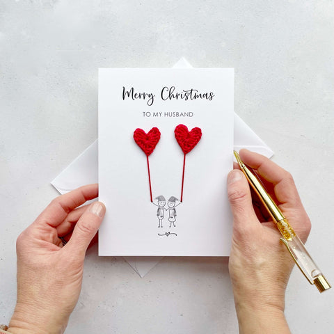 A white Christmas card featuring a line drawn couple holding hands and each holding a red crochet heart balloon. ‘Merry Christmas to my husband’ is printed at the top of the card. The couple are drawn wearing winter scarves and santa hats.