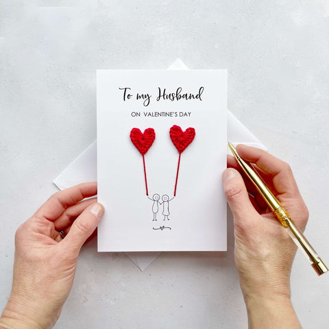 A white card featuring a line drawn couple holding hands and each holding a red crochet heart balloon.  ‘To my husband on Valentine’s Day’ is printed at the top of the card in black cursive text.