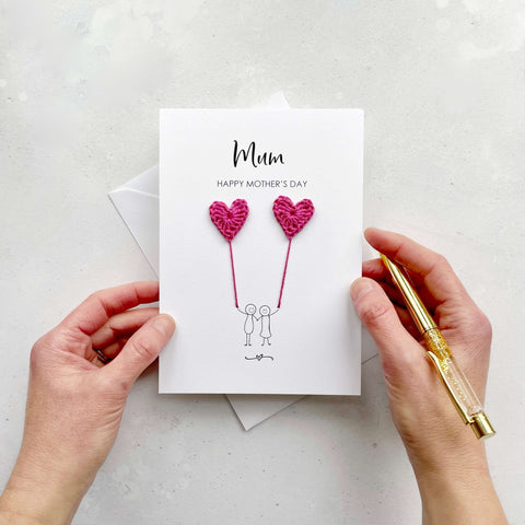 A white Mother's Day card featuring a line drawn couple holding hands and each holding a bright pink crochet heart balloon. At the top of the card, the word 'Mum' is printed in large black cursive text and just under that "Happy Mother's Day' has been printed in smaller black capitals. 