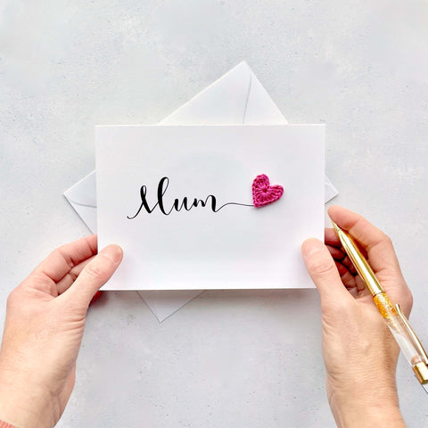 A white card with the word 'Mum' printed across the middle in black script text. There is a bright pink crochet heart stuck onto the end of the word 'Mum'