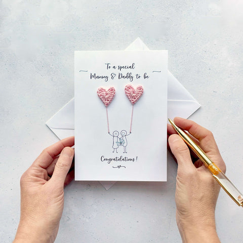 A card featuring a line drawn couple holding hands and each holding a pale pink crochet heart balloon. ‘To a special Mummy & Daddy to be’ is printed at the top of the card in black cursive text .The couple are drawn holding a baby car carrier and the word ‘Congratulations!’ is printed below the couple. 