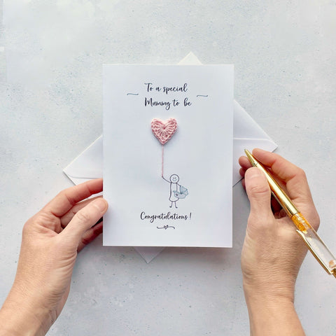A card featuring a line drawn female, holding a pale pink crochet heart balloon. ‘To a special Mummy to be’ is printed at the top of the card in black cursive text .The character  is drawn holding a baby car carrier and the word ‘Congratulations!’ is printed below her.