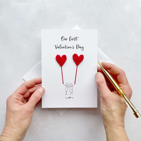 A white card featuring a line drawn couple holding hands and each holding a red crochet heart balloon.  ‘Our first Valentine’s Day’ is printed at the top of the card in black cursive text.