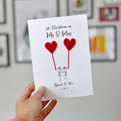 A white card featuring a line drawn couple holding hands and each holding a red crochet heart balloon. ‘1st Christmas as Mr & Mrs’ is printed at the top of the card and the couples names are printed just below the couple. The couple are drawn wearing winter scarves and santa hats.