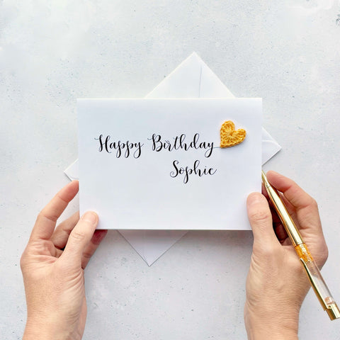 A white birthday card with 'Happy Birthday Sophie' printed in black cursice text. A yellow crochet heart has been attatched to the 'y' at the end of Birthday. 