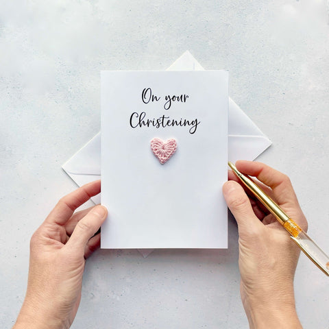 A white Christening card featuring a pale pink crochet heart. The words 'On your Christening' have been printed at the top of the card in black cursive text. 