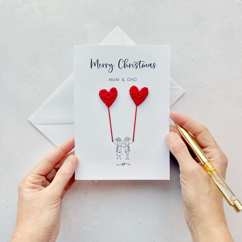 A white Christmas card featuring a line drawn couple holding hands and each holding a red crochet heart balloon. ‘Merry Christmas’ is printed at the top of the card in black cursive text and under that ‘Mum & Dad’ is printed in smaller text. The couple are drawn wearing winter scarves and santa hats.