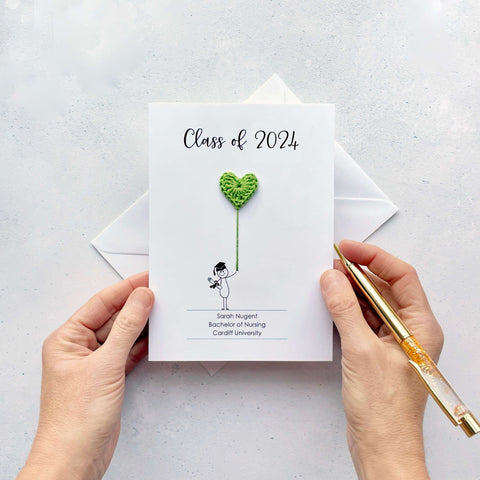 A white card with ‘Class of 2024’ printed at the top in black cursive text. There is a line drawn person holding a lime green crochet heart balloon. The person is wearing a graduation mortar board. There are 3 lines of personalisation at the bottom of the card (name/ course/ University) 