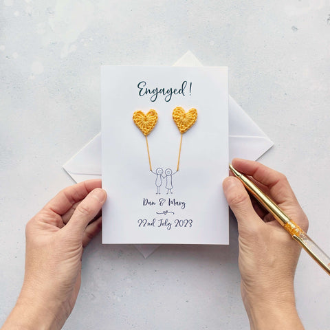 A white card featuring a line drawn couple holding hands and each holding a yellow crochet heart balloon. ‘Engaged!’ is printed at the top of the card and the couples names and date of engagement is printed just below the couple. 