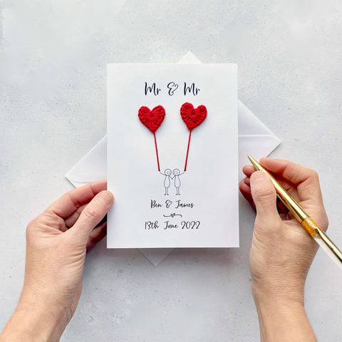 A white card featuring a line drawn couple holding hands and each holding a red crochet heart balloon. ‘Mr & Mr’ is printed at the top of the card and the couples names and wedding date is printed just below the couple. 