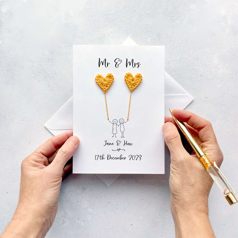 A white card featuring a line drawn couple holding hands and each holding a yellow crochet heart balloon. ‘Mr & Mrs’ is printed at the top of the card and the couples names and wedding date is printed just below the couple. 