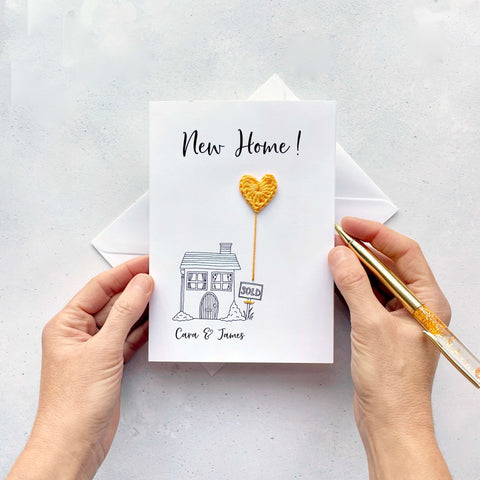 A white card with line drawn house on the front. A yellow crochet heart has been attatched to the card to look like  a heart shaped balloonn coming up from the chimney. ’New Home!’ is printed at the top of the card in black cursive text. The couples names have been printed below the house.  