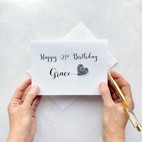 A white birthday card with 'Happy 21st Birthday Grace' printed on it in blackk cursive text. The words Grace appears larger than the rest of the text and there is a grey crochet heart at the end of her name. 