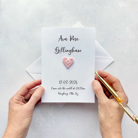 A white personalised new baby card that features a pale pnk crochet heart. The babies name, date of birth, weight and time of birth are all printed in black cursive text. A lovely keepsake. 