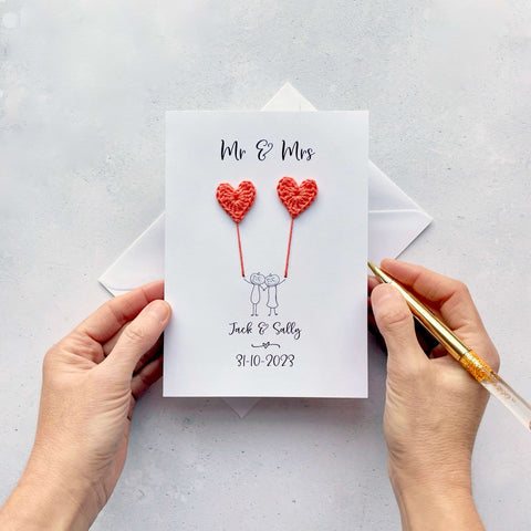 A white card featuring a line drawn couple holding hands and each holding a yellow crochet heart balloon. ‘Mr & Mrs’ is printed at the top of the card and the couples names and wedding date is printed just below the couple. The couple have pumkins as heads and heart shaped cut out eyes. 