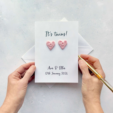 A white personalised new twins card featuring two pale pink crochet hearts. 'It's twins!' is printed at the top of the card in black cursive text and the babies names and date of birth are printed at the bottom of the card. 