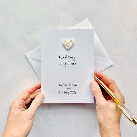 A white greetings card with 'Wedding acceotance' printed in the center in black script front. Above this text is an ivory coloured cotton crochet heart. The bride & grooms name and wedding date is printed at the botton of the card. 